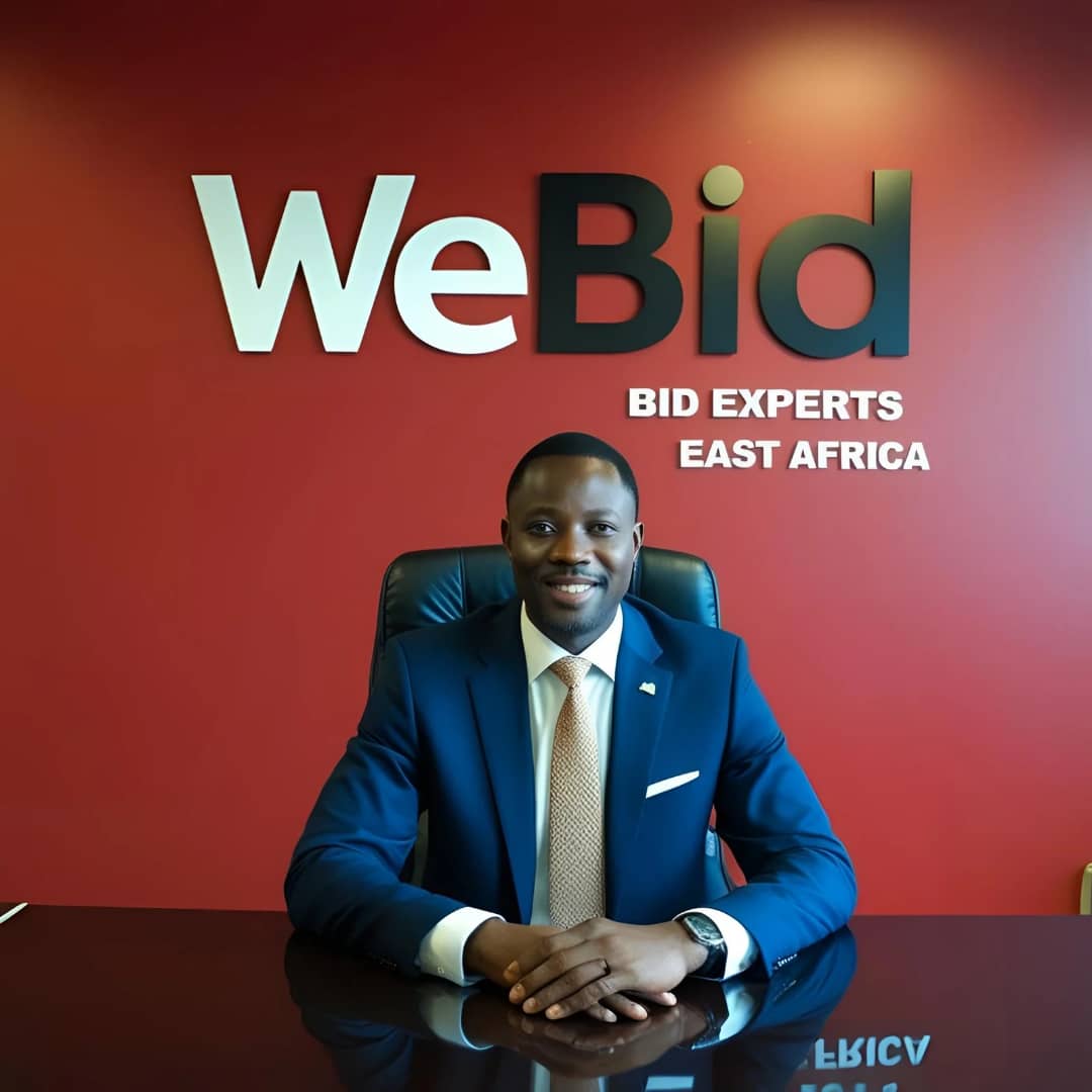 Bid writing expert in a blue suit sits at a desk in an office chair, with hands clasped. Behind them is a red wall with the text "WeBid" in large white and black letters, and "BID EXPERTS EAST AFRICA" in smaller white letters. The desk has a reflective surface showing part of the background text.